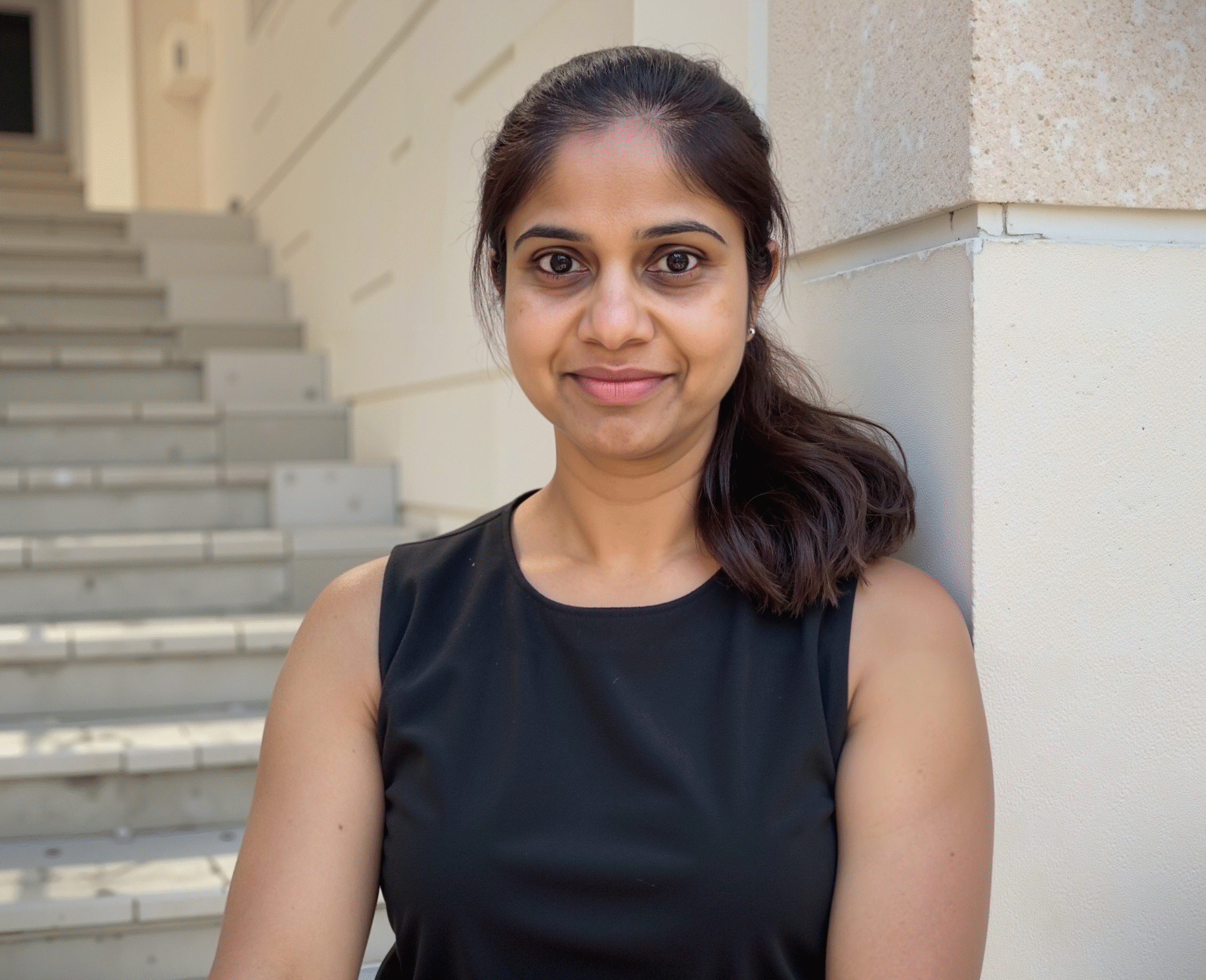 Portrait of a woman in a sleeveless black top standing by a staircase and smiling at the camera.