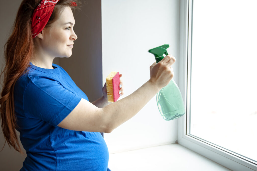 Pregnant woman in a blue shirt and red headband spraying cleaner on a window while holding a sponge.